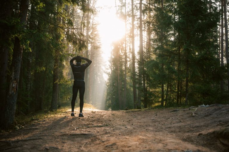 Žmogus sportine apranga stovi ant miško kelio, rankas uždėjęs ant galvos, pro pušis šviečia saulė